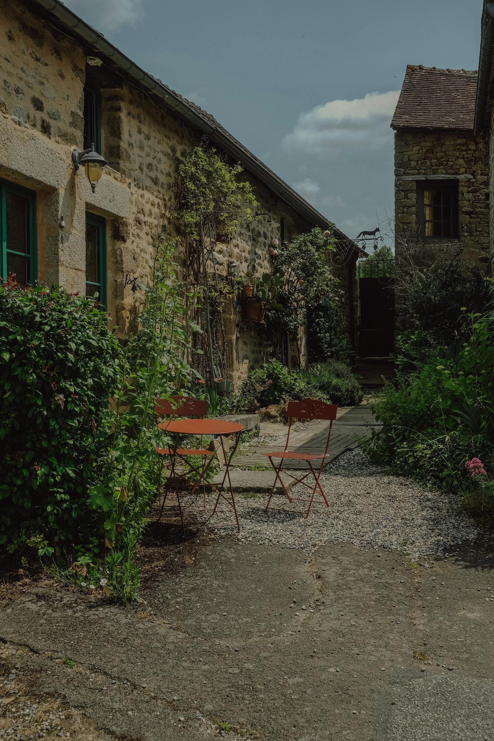 A quaint alley in a rustic setting featuring outdoor red chairs and table surrounded by greenery.